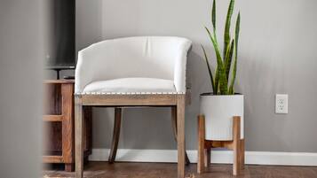 Una silla tapizada de blanco con patas de madera, una planta en maceta con hojas verdes largas y un televisor sobre un mueble de madera.