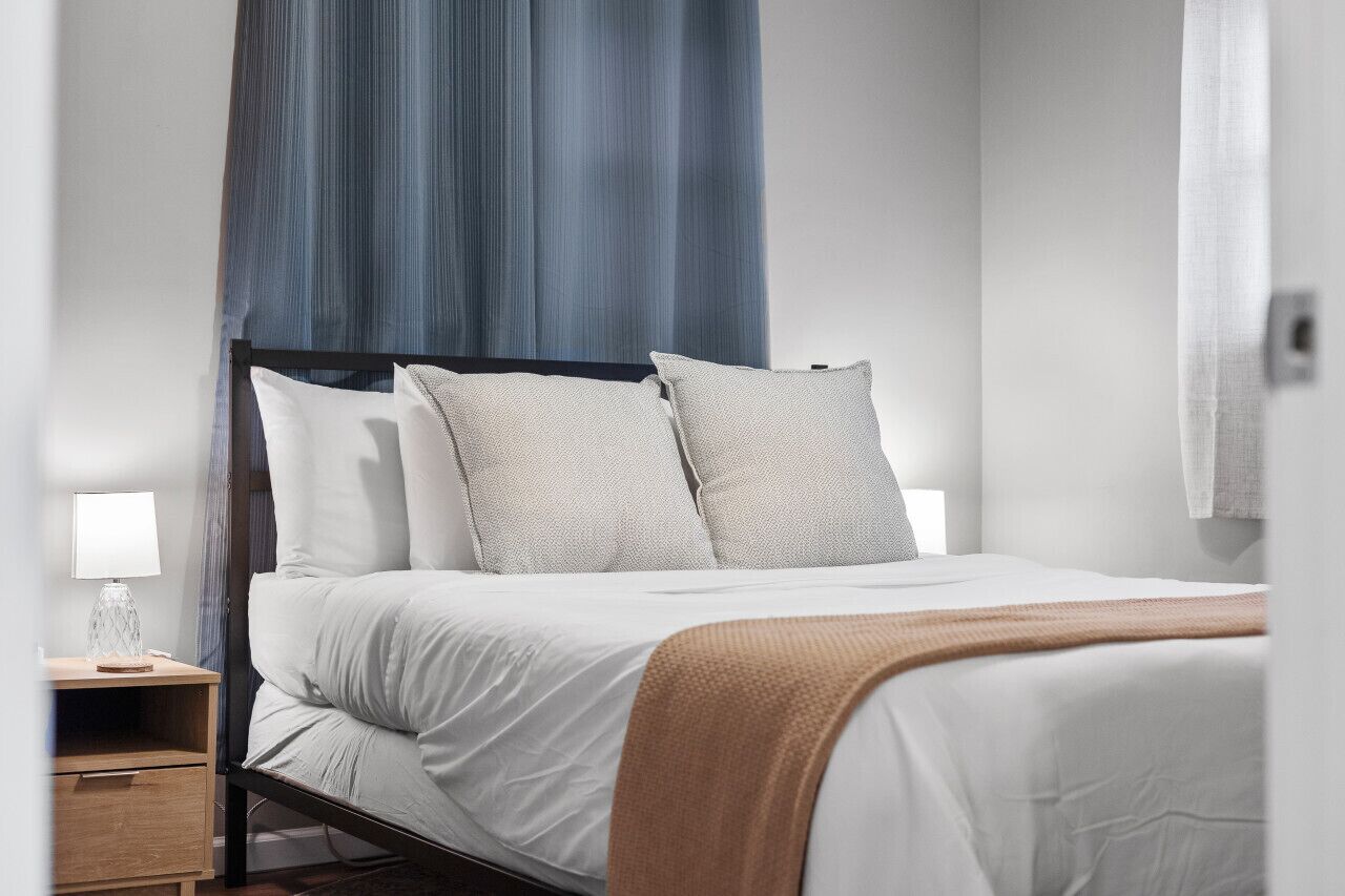 Een net opgemaakt bed met witte lakens en twee kussens, een nachtkastje met een lamp, en een blauw gordijn.