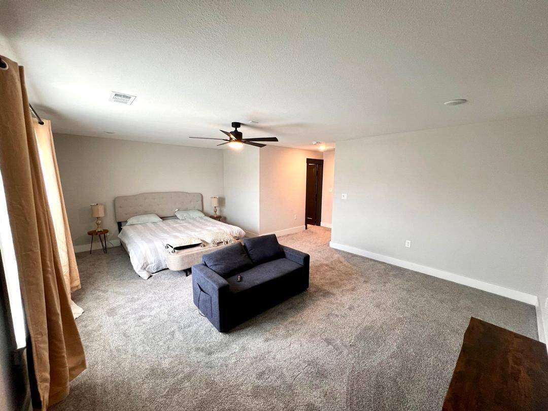 Un dormitorio con cama, un sofá oscuro, un ventilador de techo y una puerta que da a otra habitación.