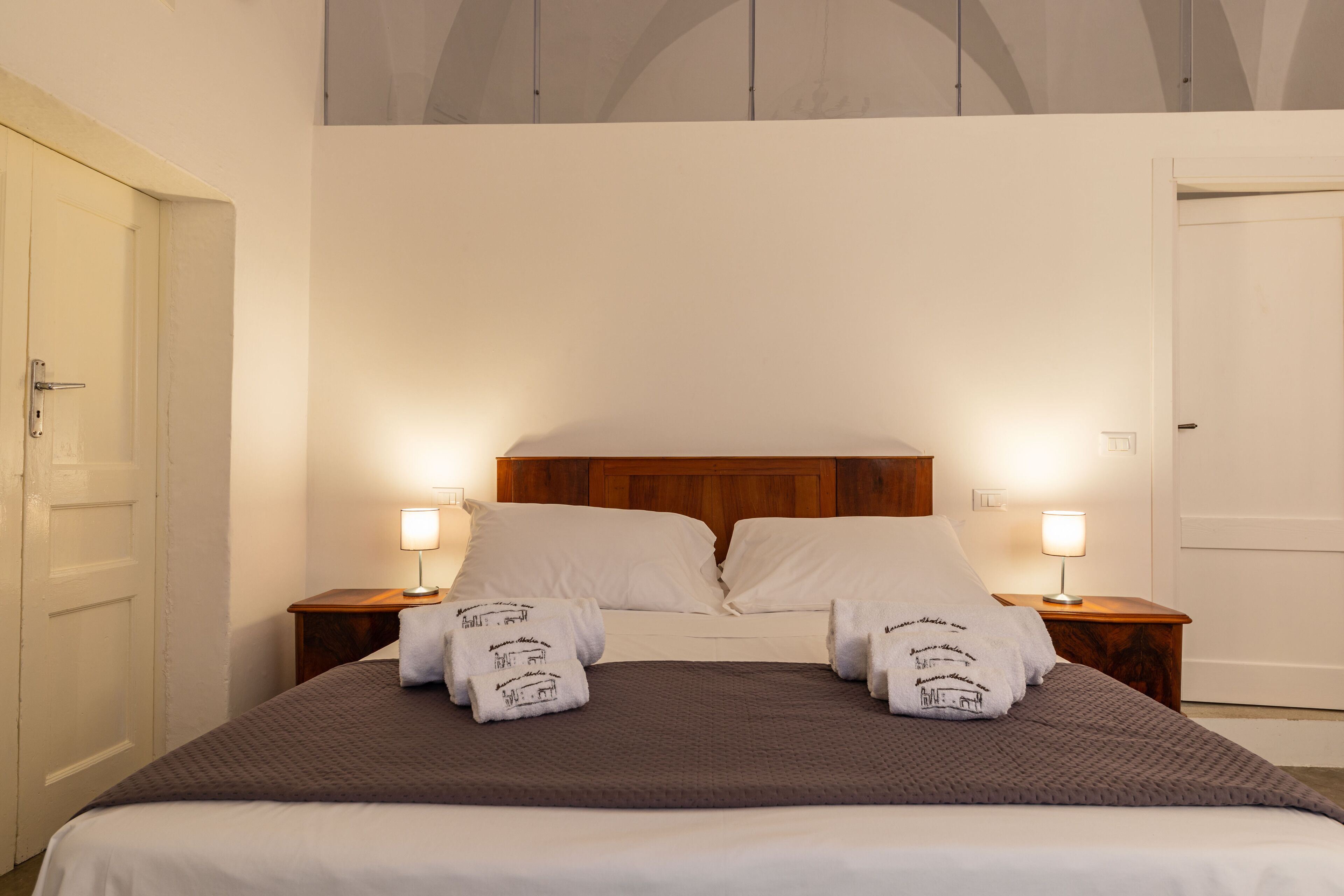 Ein ordentlich gemachtes Bett mit weißen Bettwäsche und zwei Kissen, das von Nachttischen mit Lampen gesäumt wird, in einem Raum mit schlichten Wänden und einer Tür links.