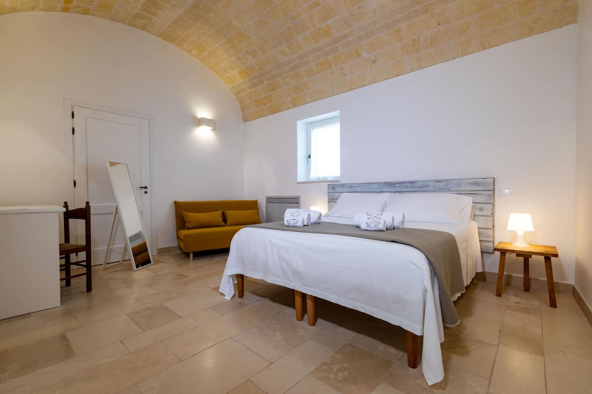 A bedroom with a bed, a window, a sofa, a lamp, and a door.
