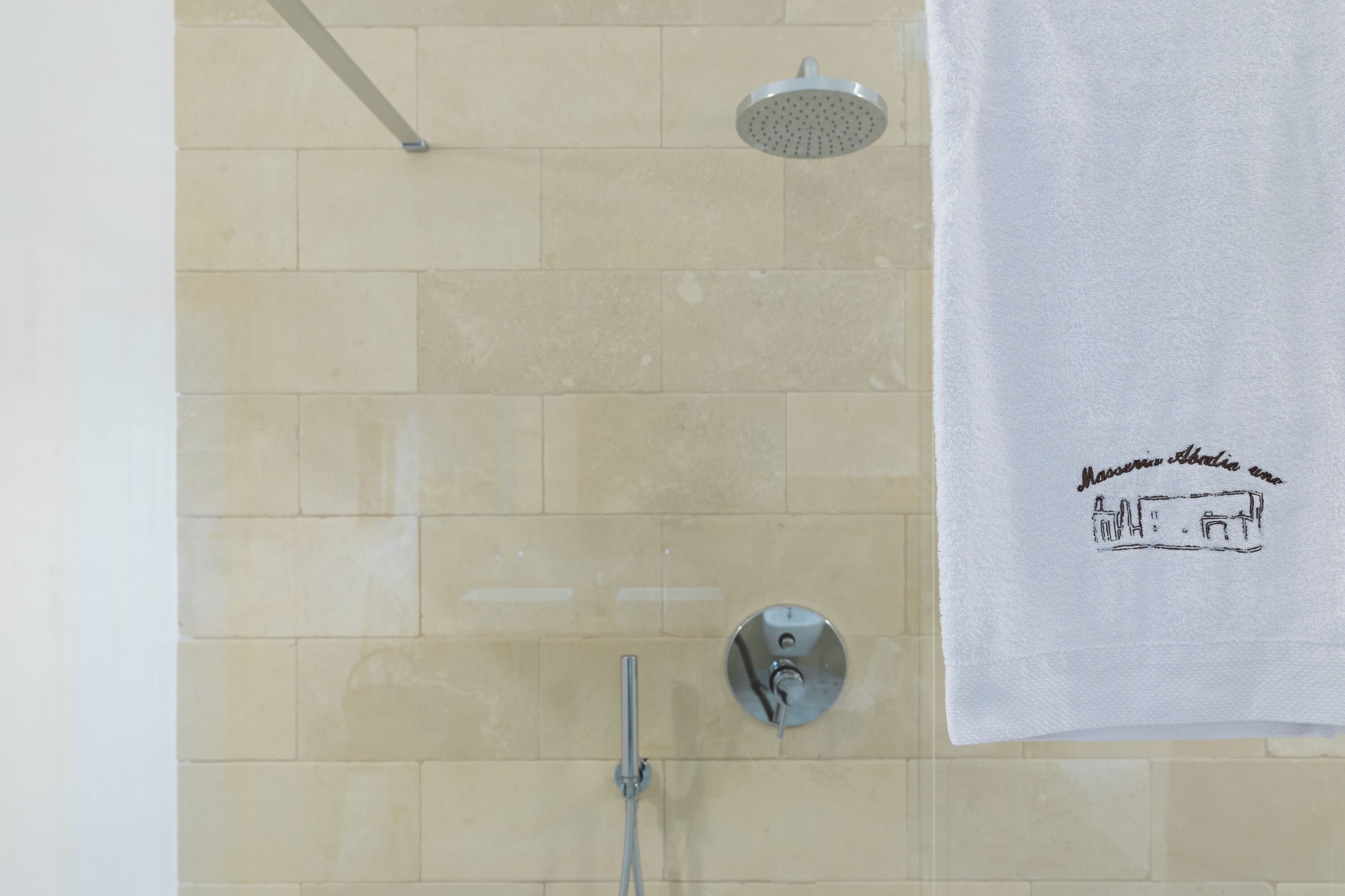 Une salle de bain avec des carreaux de couleur beige, une pomme de douche, un pommeau de douche amovible et une serviette ornée d’une illustration représentant un bâtiment.