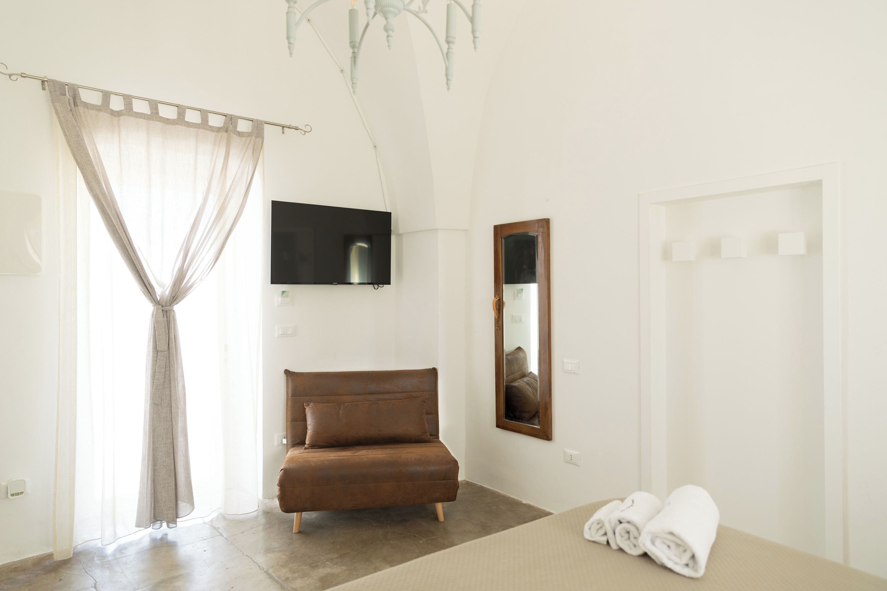 Un dormitorio con un sofá marrón, un televisor de pantalla plana, un espejo y una cama con toallas enrolladas.