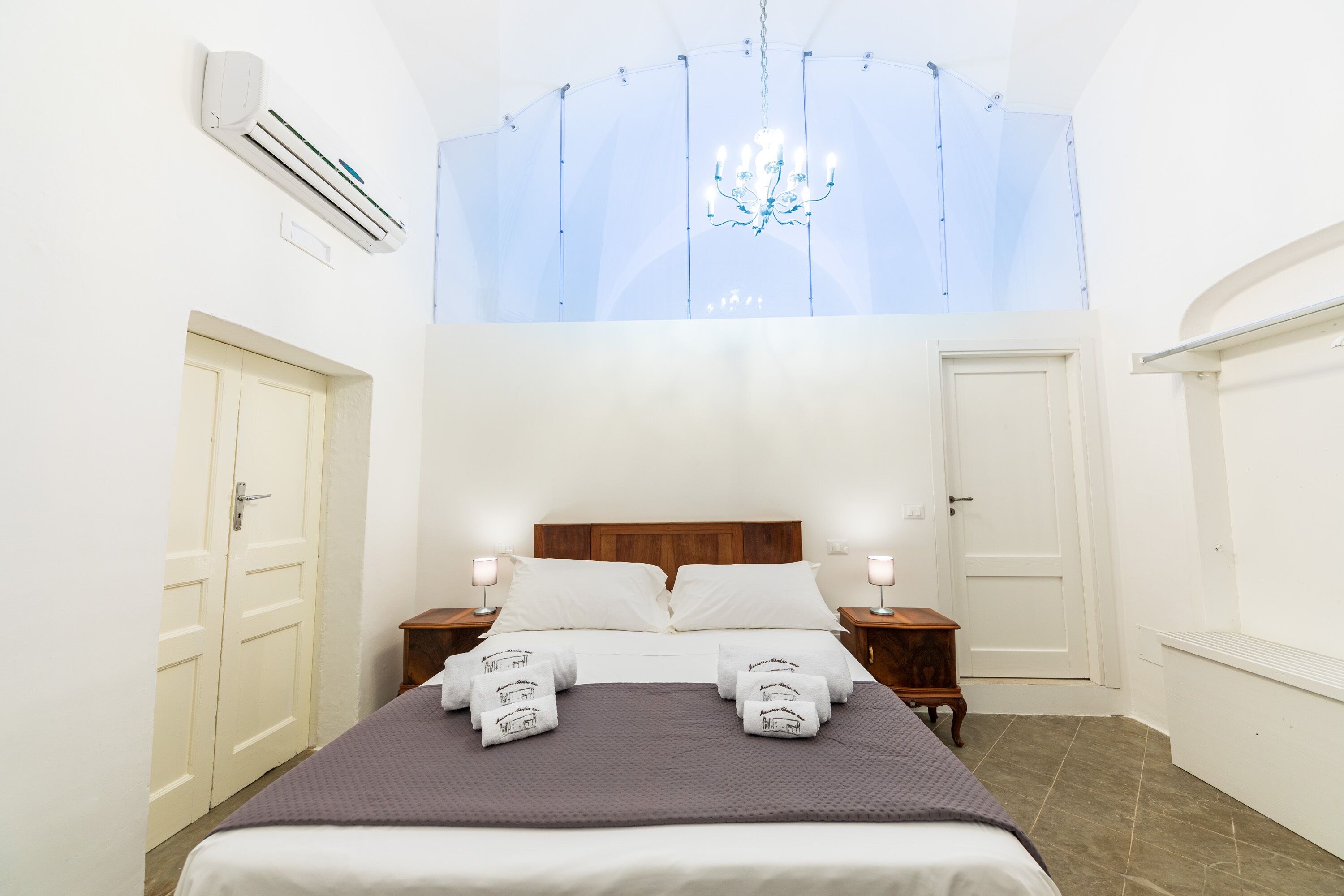 Habitación de hotel con cama, dos mesitas de noche, una araña y ventana con vistas.