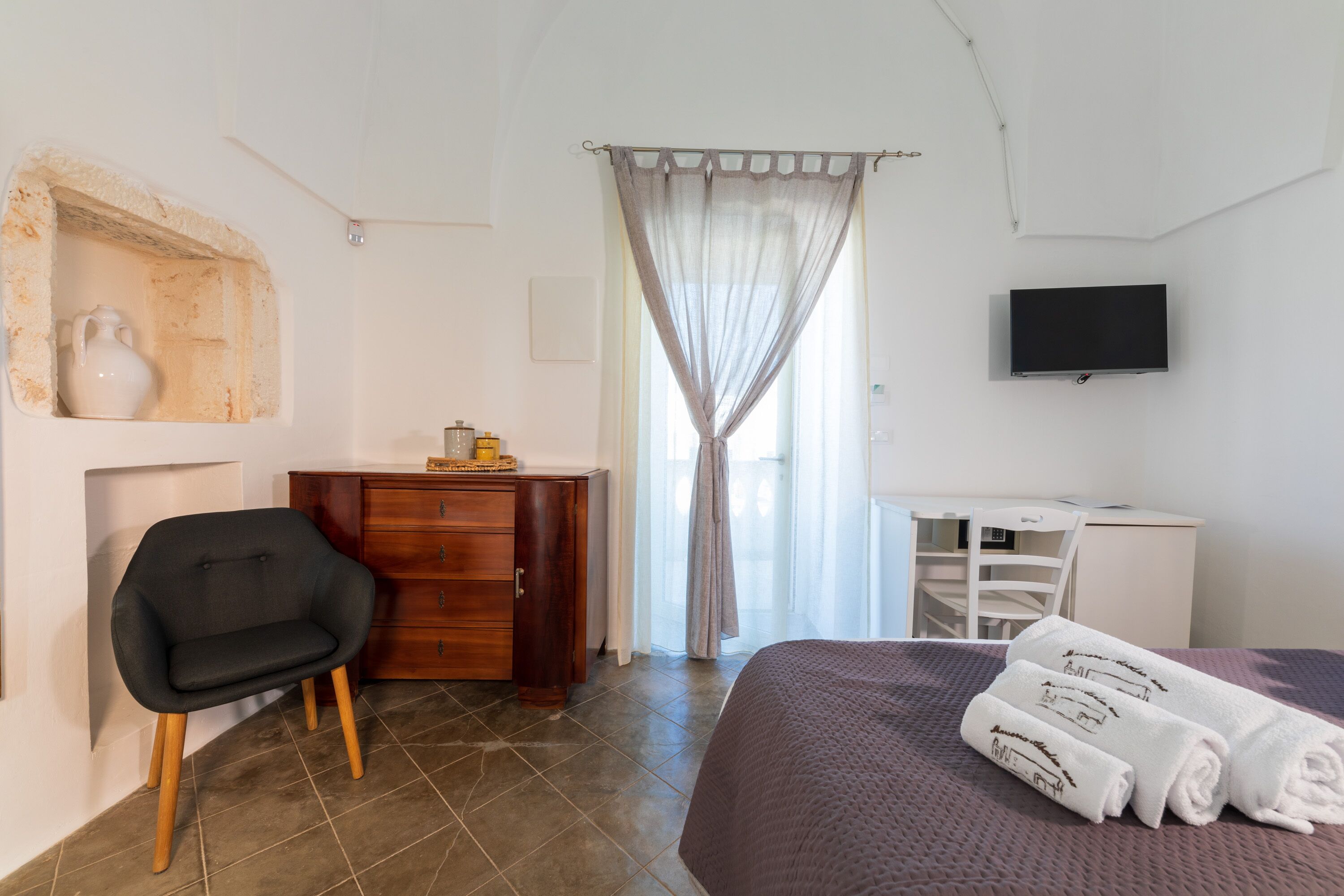 Une chambre à coucher comprenant un lit, un bureau, une chaise, une commode, un téléviseur et une fenêtre avec des rideaux.