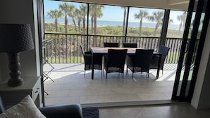 A balcony with a table and chairs, a ceiling fan, and a view of the beach and palm trees.