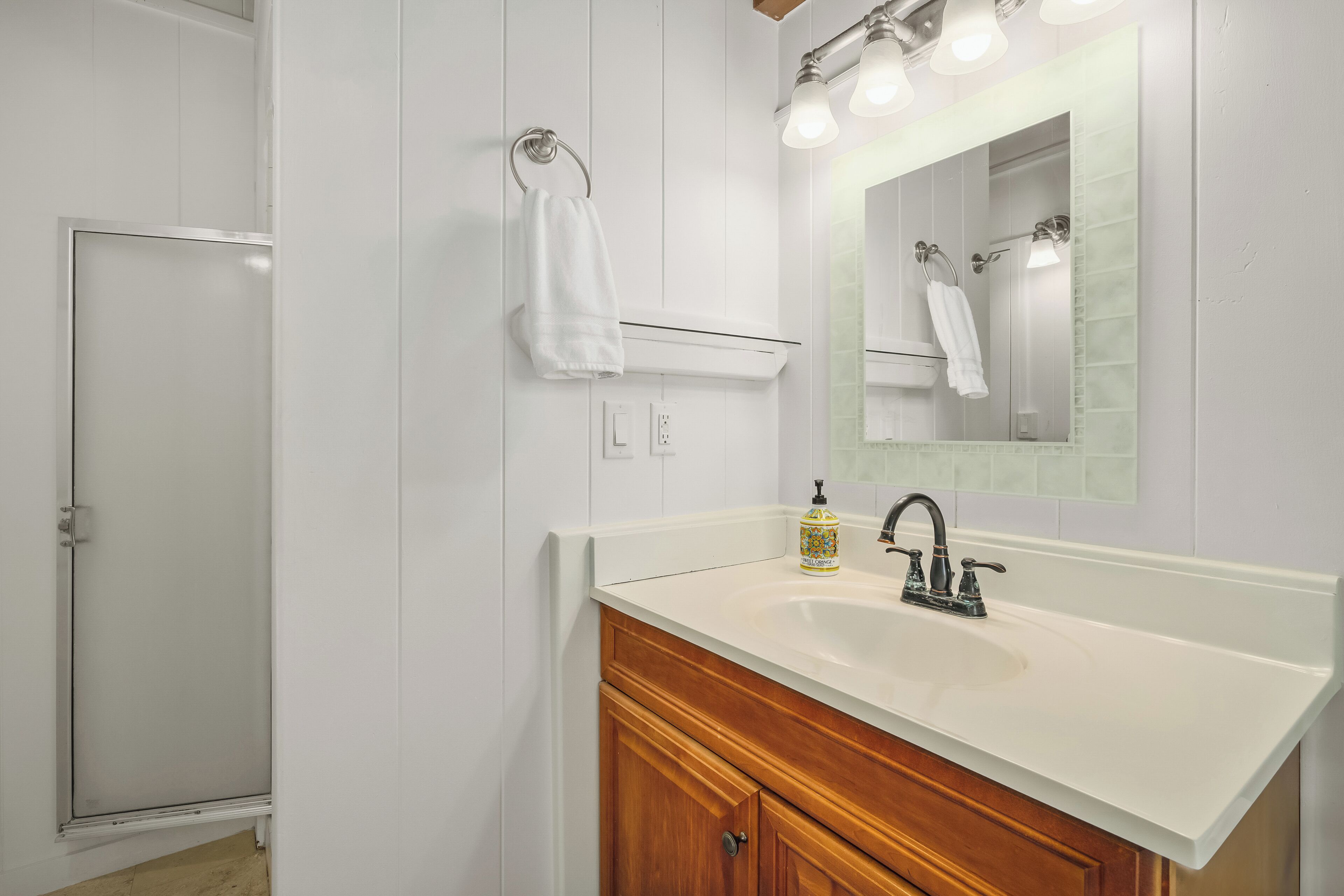 A bathroom with a white sink, wooden vanity, a glass shower enclosure, and a mirror with lights.