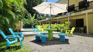 A covered outdoor seating area with chairs and tables, surrounded by lush greenery and a building with balconies. - Family Hotel (Cahuita)