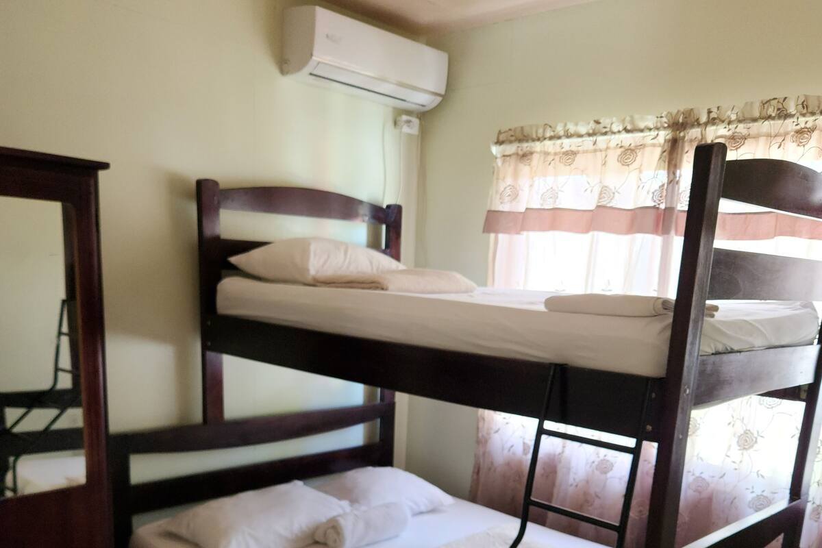 A bunk bed with a wooden frame, a white mattress, and a pillow on the top bunk. There is a small wooden table beside the bed, and an air conditioning unit mounted on the wall above the bed.