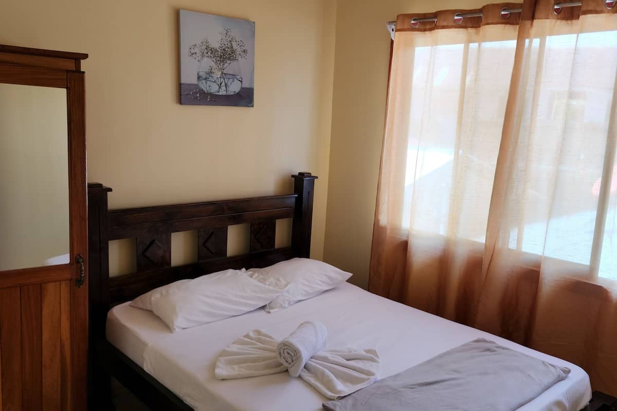 A neatly made bed with white linens and towels, a wooden headboard, a small painting on the wall, and a window with sheer curtains.