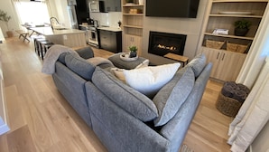 A modern living room with a grey sectional sofa, a flat-screen TV mounted above a fireplace, and a kitchen area with dark cabinetry and a microwave. - Carsonland Getaways Panorama BC (Panorama)