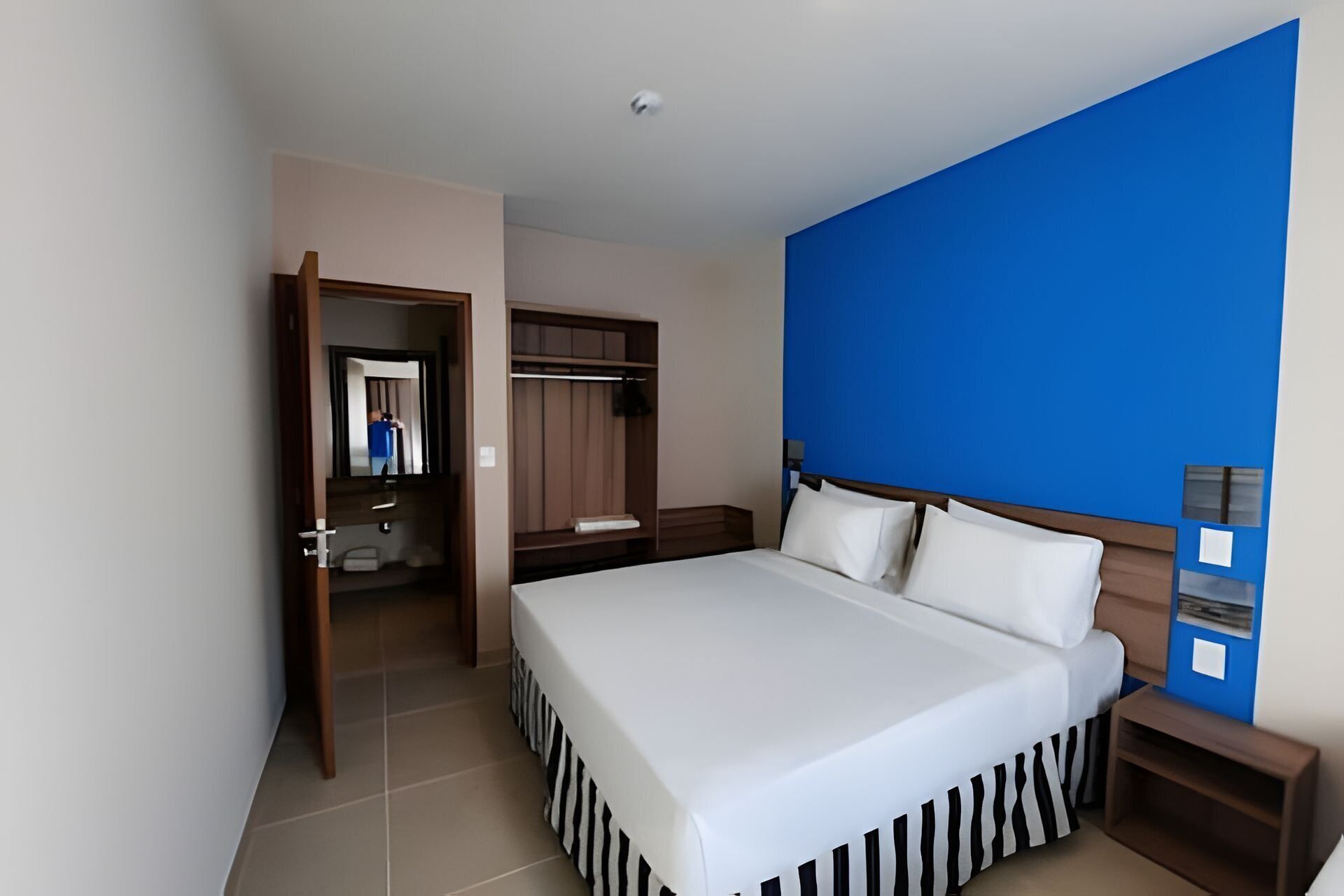 Quarto de hotel com uma cama grande, um guarda-roupa, um espelho e uma parede azul decorativa.