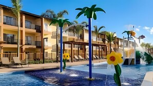 A swimming pool with water features, including a fountain and flower-shaped structures, surrounded by a building with balconies and a poolside area. - Ondas Praia Resort (Porto Seguro)