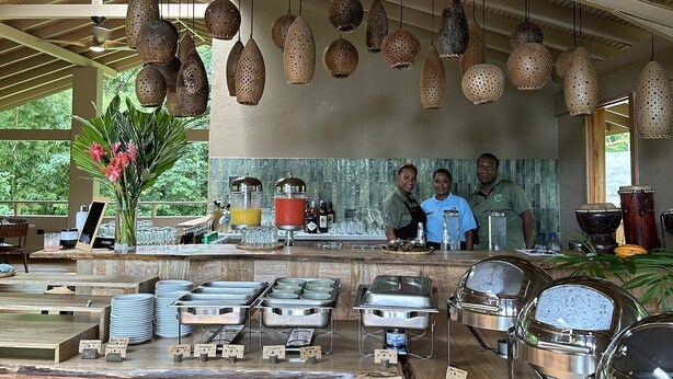 A buffet setup with various food stations, hanging pendant lights, and a view of the outdoors. - HADCO Experiences at AWNC (Lopinot)