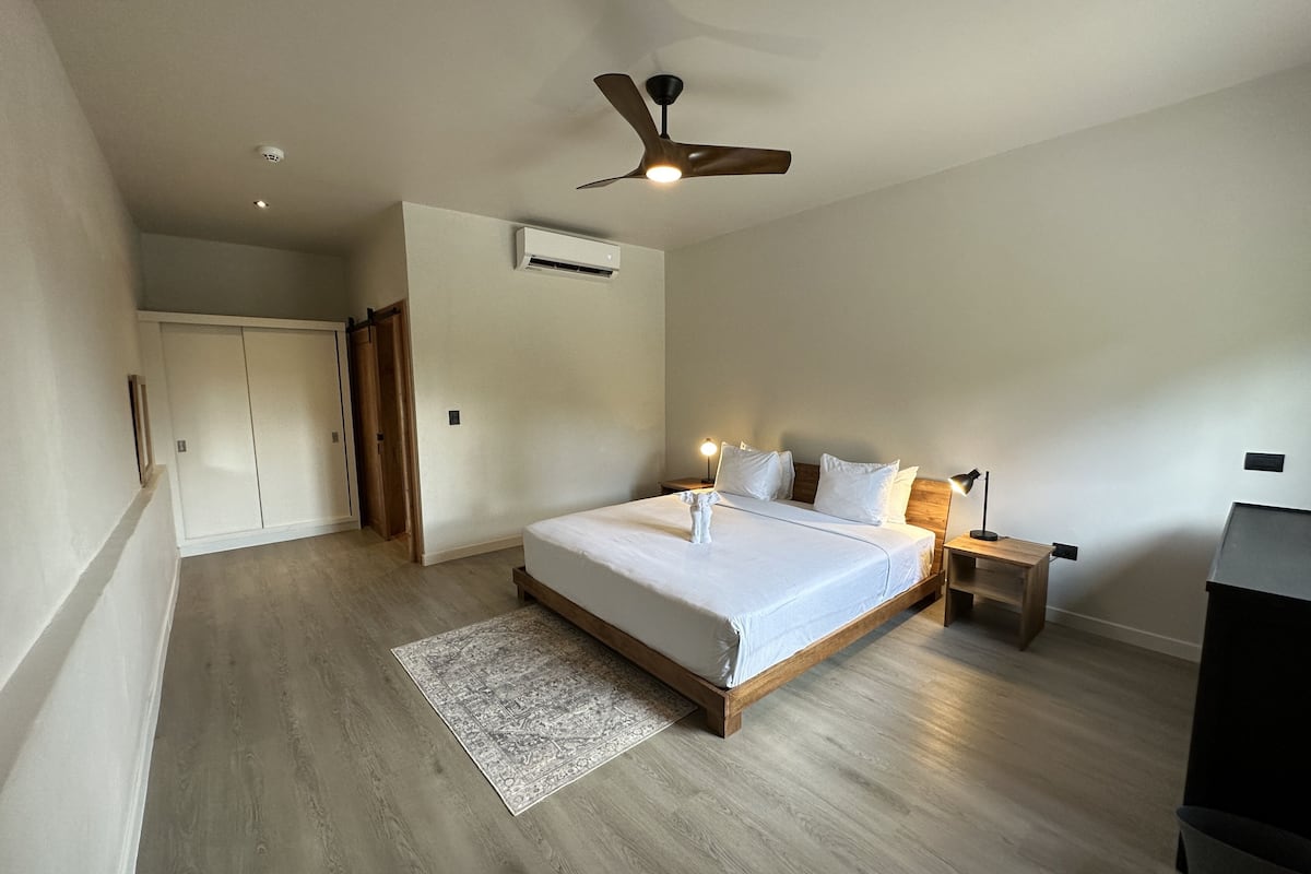 Une chambre à coucher avec un lit, des lampes de chevet, un ventilateur de plafond, un appareil de climatisation et un plancher en bois.