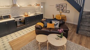 A modern kitchen with dark cabinets, a built-in oven, and a microwave. There is a dining area with a brown leather sofa and two round tables. A staircase with a blue railing leads to another level. - The Hare (Nantwich)