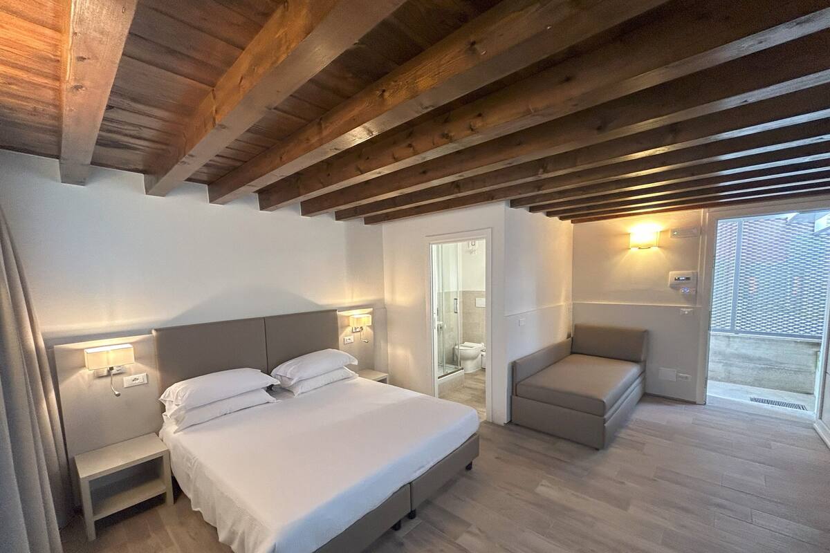 A bedroom with a bed, bedside tables, a nightstand, a seating area with a chair, and a view of the outside.