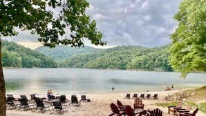 A lakeside setting with wooden chairs and tables arranged on the sand, overlooking a calm lake and forested hills. - Amazing views at L’Oursière in Bear Lake Reserve, from the new  hot tub. (Tuckasegee)