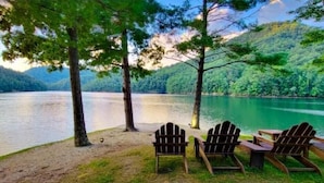 A lakeside setting with wooden chairs and a table, surrounded by trees and a view of a lake. - Amazing views at L’Oursière in Bear Lake Reserve, from the new  hot tub. (Tuckasegee)