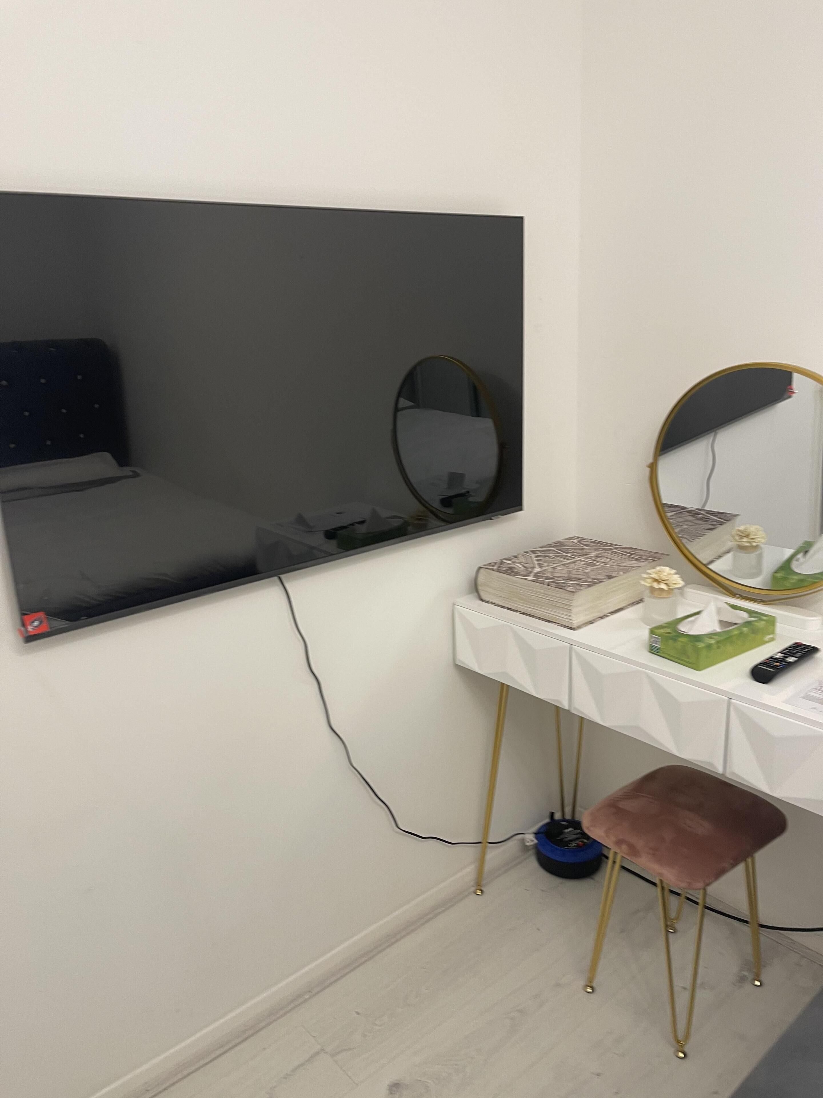 A room with a white vanity, a large mirror, a flat-screen TV mounted on the wall, and a small stool.