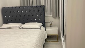 A modern bedroom with a tufted headboard, a white bedspread, a bedside table with a lamp, and a window with curtains. - Deptford - 1 Bed - 1 Bathroom - Parking (London)