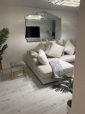 A modern living room with a white sofa, a small wooden stool, and a large mirror with a decorative frame. - Deptford - 1 Bed - 1 Bathroom - Parking (London)