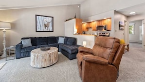 Ein modernes Wohnzimmer mit einem Ecksofa, einem runden Couchtisch und einem Ledersessel. Im Hintergrund ist eine Küche mit Holzausstattung und einer Mikrowelle erkennbar.