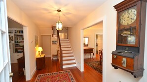 A hallway with a wooden staircase, a large wall clock, and a patterned rug.