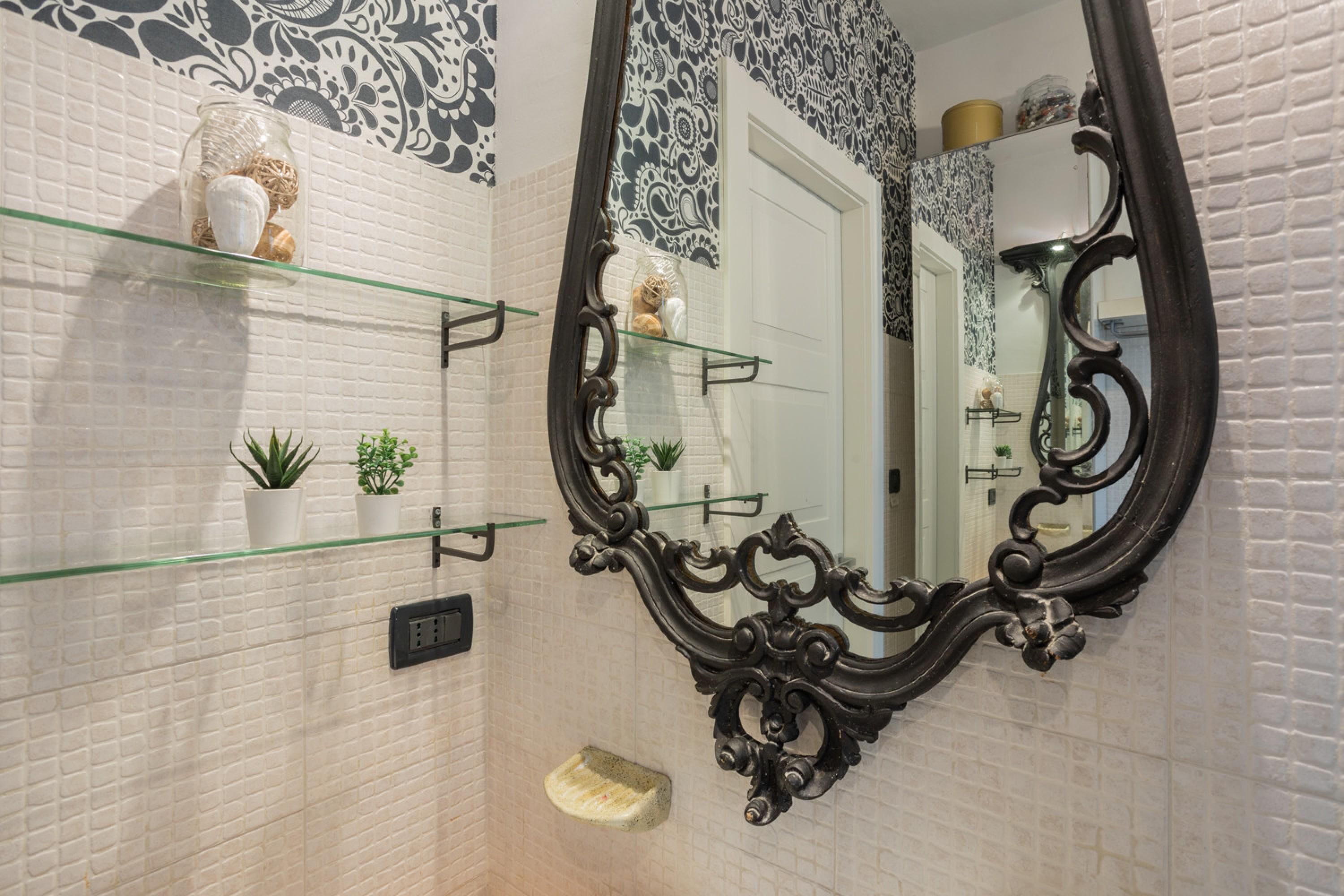 A bathroom with a white tiled wall, a glass shelf with decorative items, a black ornate mirror, and a small step for accessibility.