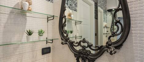 A bathroom with a white tiled wall, a glass shelf with decorative items, a black ornate mirror, and a small step for accessibility.