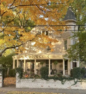 A large, historic house with a prominent tower, a covered porch, and a brick wall with greenery.