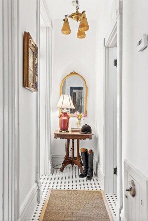 A narrow hallway with a white door, a mirror with a gold frame, a table with a lamp, a vase, and a pair of boots. - Historic Ward Home - Experience history in the horse country! 5br 5.5bth (Lexington)