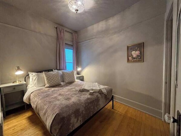 A bedroom with a bed, two bedside tables, a nightstand, a window with curtains, a ceiling light, and a framed picture on the wall.