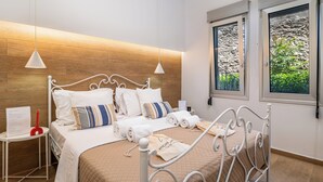 Een netjes opgemaakt bed met witte lakens en decoratieve kussens, een nachtkastje met een rood object, en een raam met uitzicht op groen.