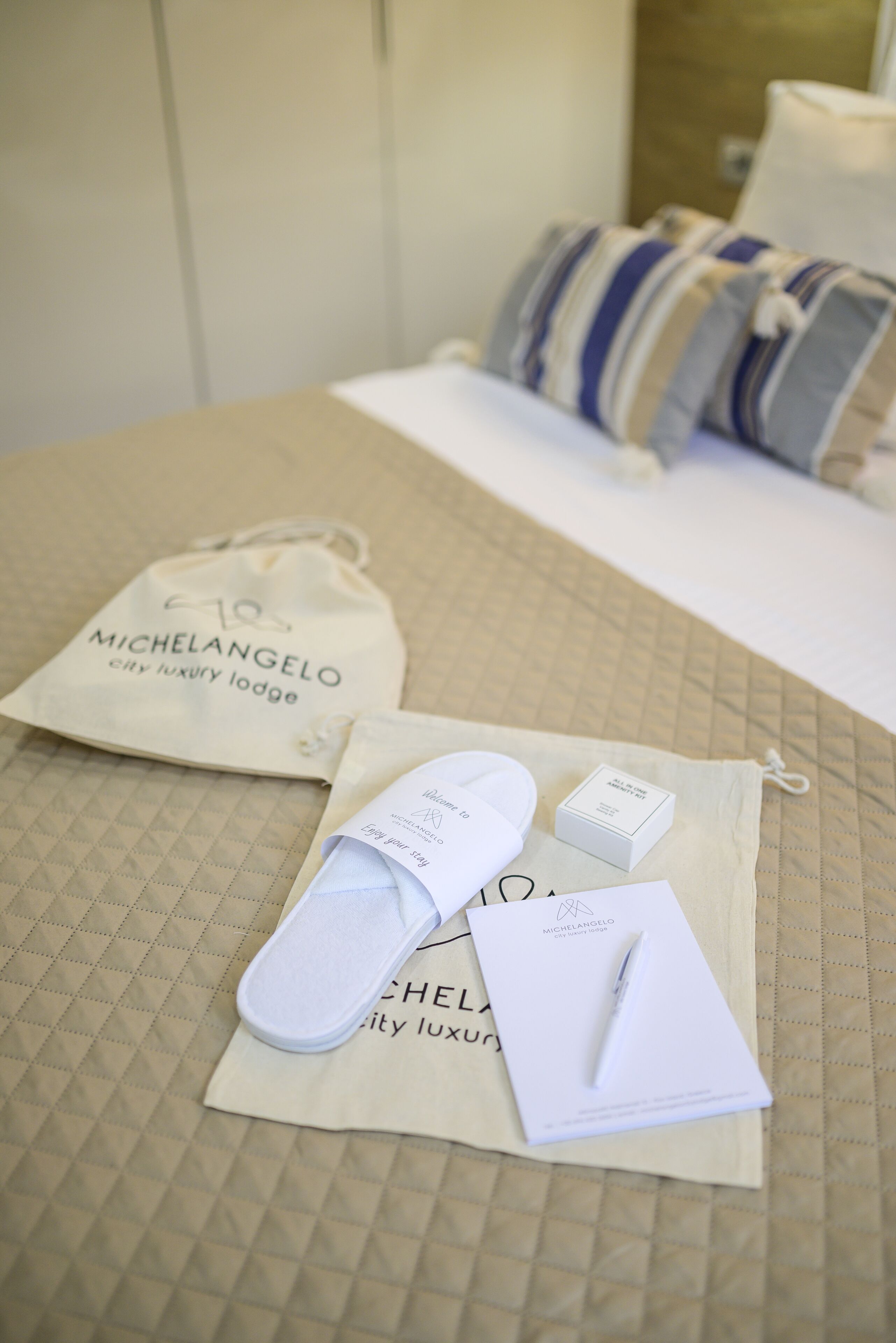 Una cama con cubierta acolchada, una almohada a rayas, una almohada blanca, una bolsa blanca y una caja blanca sobre una superficie color beige.