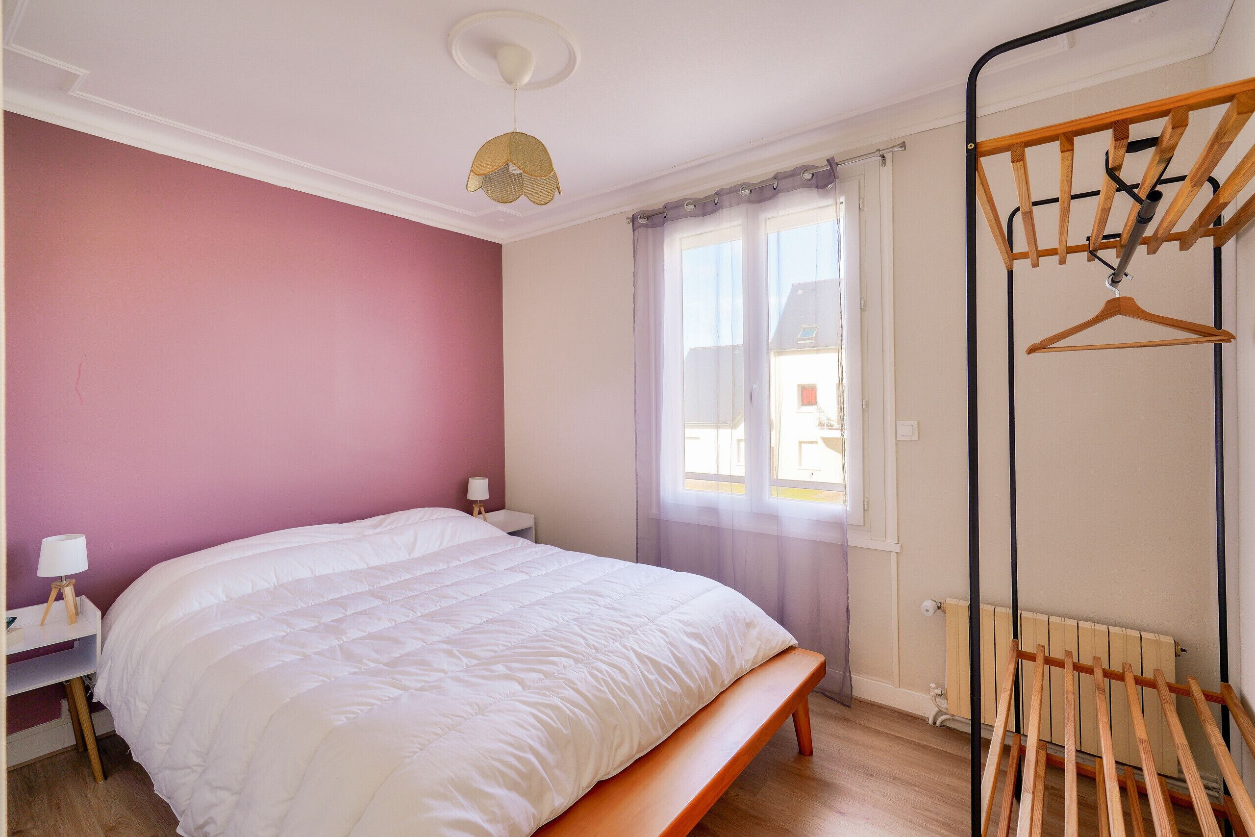 Een slaapkamer met een bed, nachtkastje, lamp, raam met gordijnen en een houten wasrek.