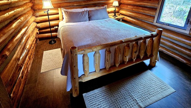 A log cabin bedroom with a wooden bed, two bedside lamps, and a window with a view of the outdoors. - Cozy log cabin with hot tub in Markleysburg! (Markleysburg)