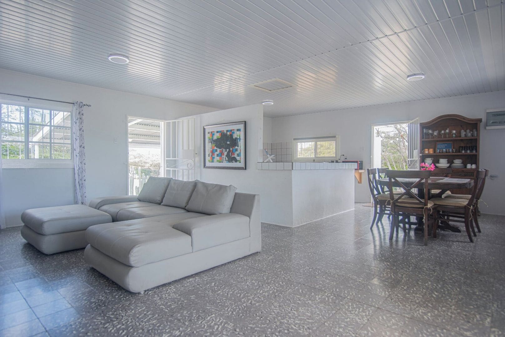 A modern living room with a sectional sofa, a dining area with wooden chairs, and a kitchen with a white countertop.