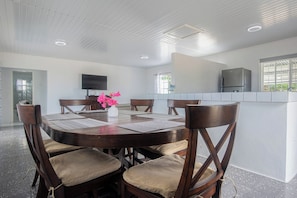 A modern dining area with a round table, cushioned chairs, a vase with flowers, and a kitchen in the background. - Aruban Home Close to Beach (Oranjestad)