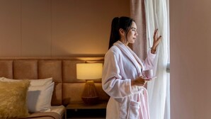 Una persona in accappatoio in piedi vicino a una finestra in una camera d'albergo, con un letto, un comodino e una lampada.