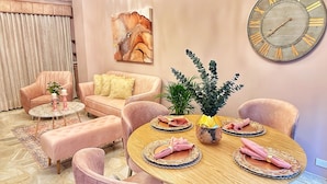 Un soggiorno moderno con un tavolo da pranzo rotondo in legno apparecchiato per quattro persone, circondato da poltrone rosa e un divano. Si notano anche una grande sveglia a parete e un quadro.