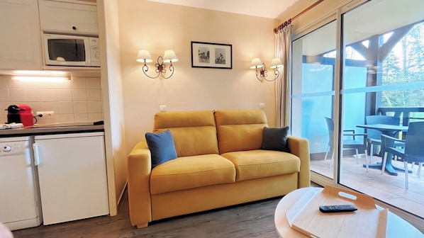 A yellow sofa with two pillows, a kitchen area with a microwave, a red coffee machine, and a dining area with a table and chairs.