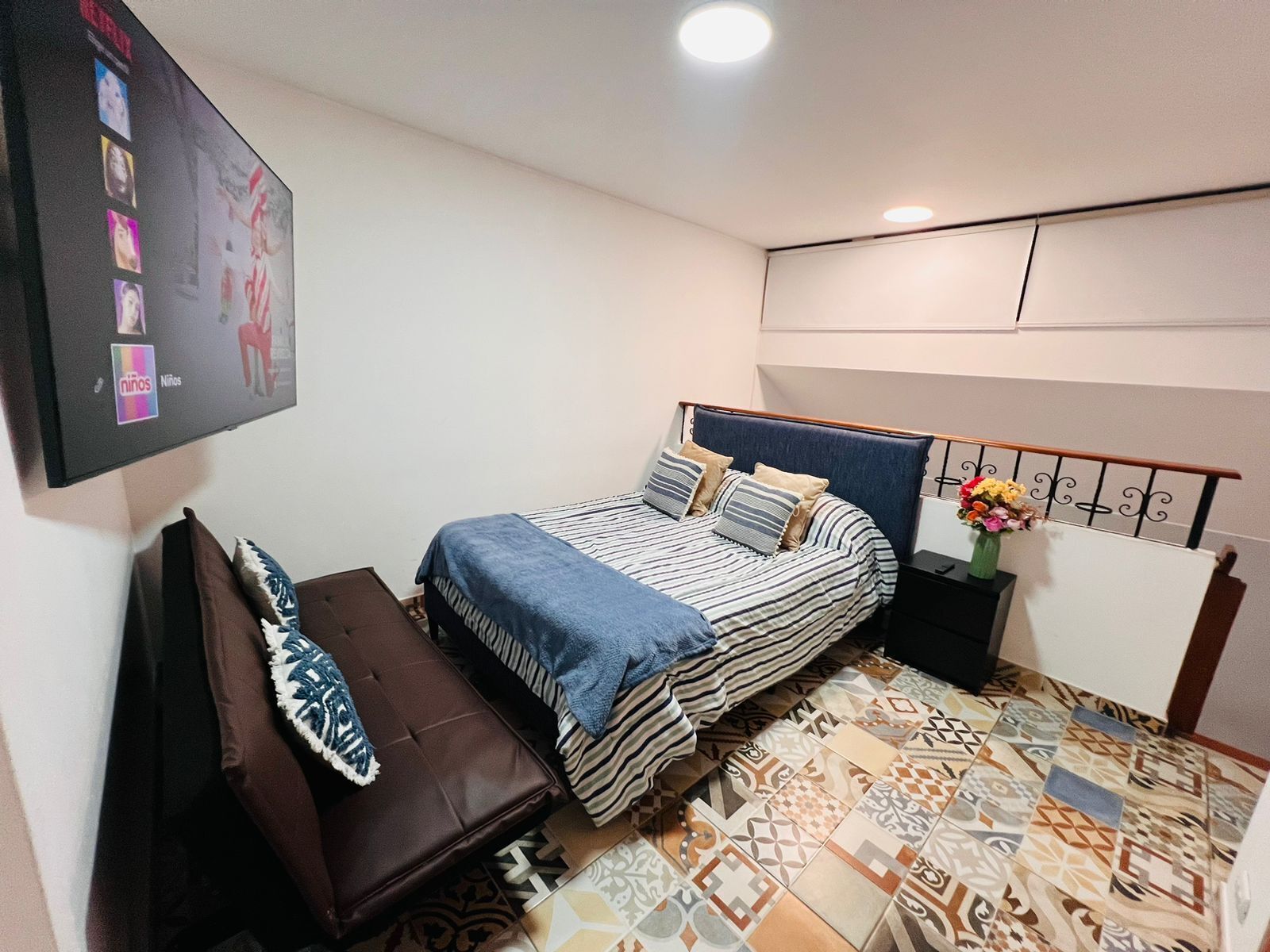 Un dormitorio con cama, mesita de noche, banco y televisor montado en la pared.