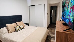A modern bedroom with a large bed, a TV on a wooden stand, and a door leading to another room. - Tezza Polo Aparts (Santiago de Surco)