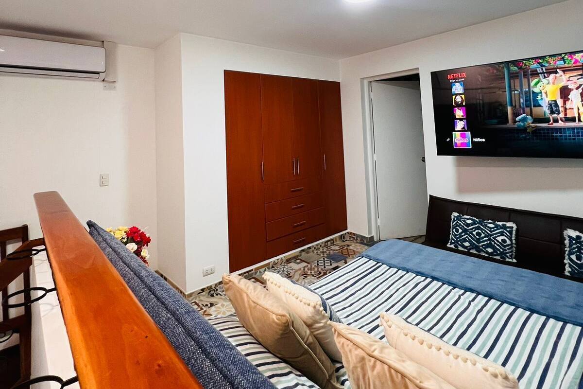 A bedroom with a bed, a wooden wardrobe, a television mounted on the wall, and an air conditioning unit on the wall.
