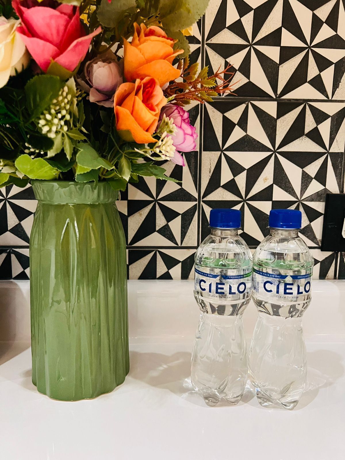 Un jarrón verde con flores y dos botellas de agua con la etiqueta 'Cielo' sobre una superficie blanca, frente a un salpicadero de baldosas geométricas en blanco y negro.