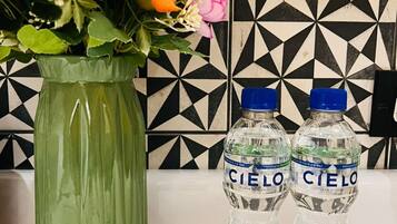 Un jarrón verde con flores y dos botellas de agua con la etiqueta 'Cielo' sobre una superficie blanca, frente a un salpicadero de baldosas geométricas en blanco y negro.