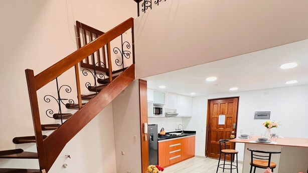 A modern living room with a wooden staircase, a red sofa, a blue armchair, and a coffee table with a vase of flowers. - Tezza Polo Aparts (Santiago de Surco)