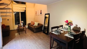 A living room with a sofa, a dining table with chairs, a small table with a vase, and a window with a curtain. - Tezza Polo Aparts (Santiago de Surco)