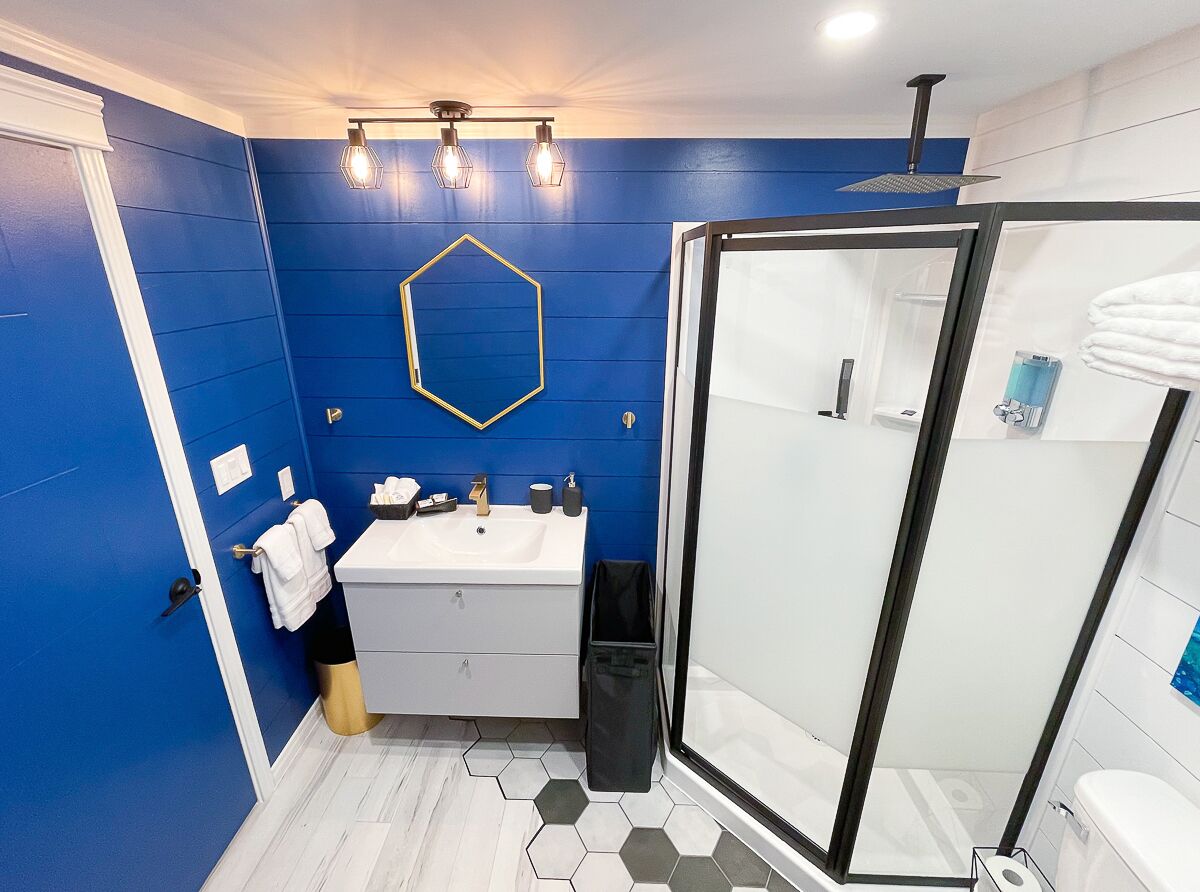 Een badkamer met een blauwe wand, een witte wastafel, een douchecabine van glas en een zeshoekige spiegel.